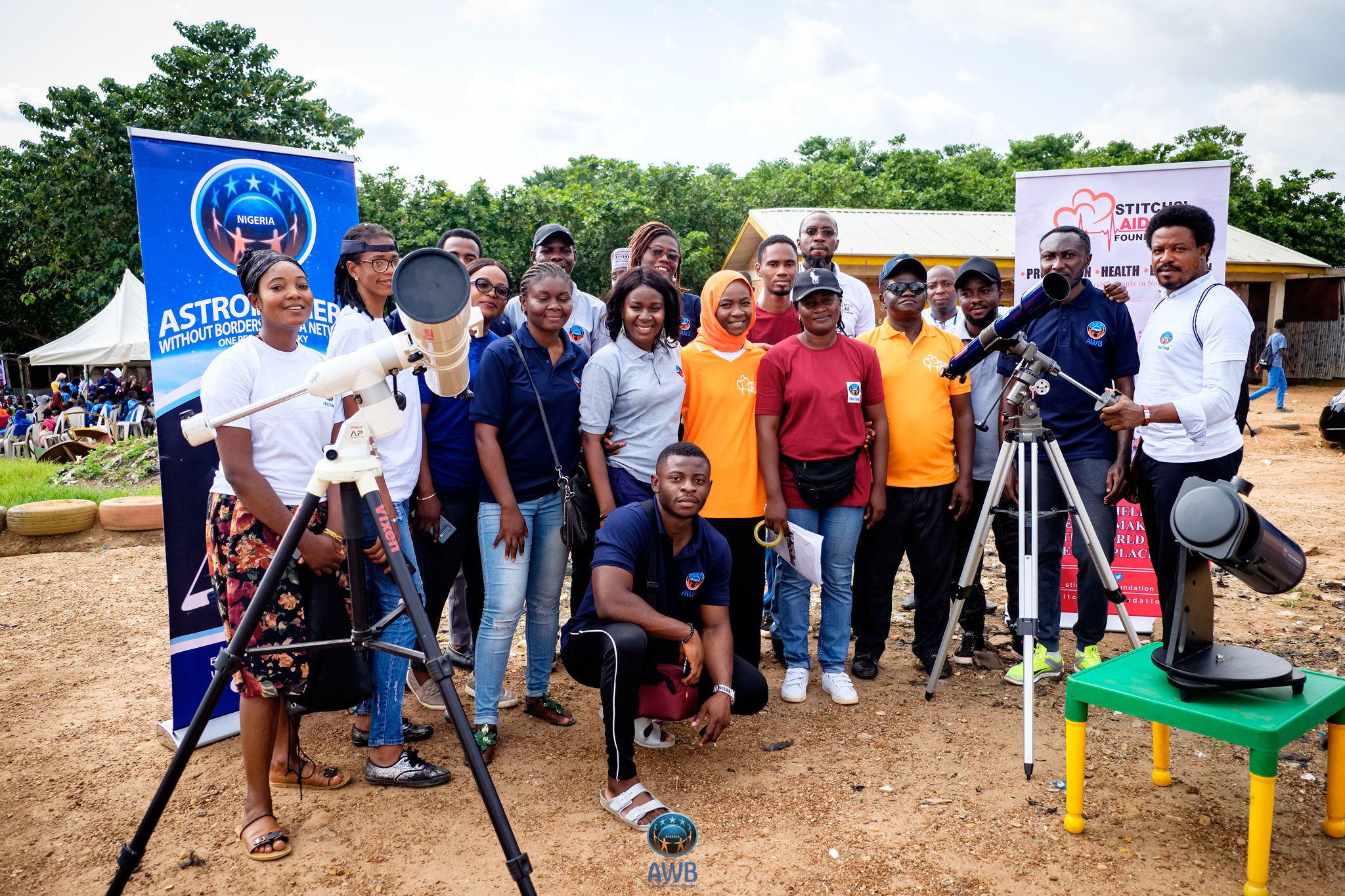 Seventeen dark-skinned adults smile for a photo at a dirt camp with tents, signs, and some grass. There are three telescopes.